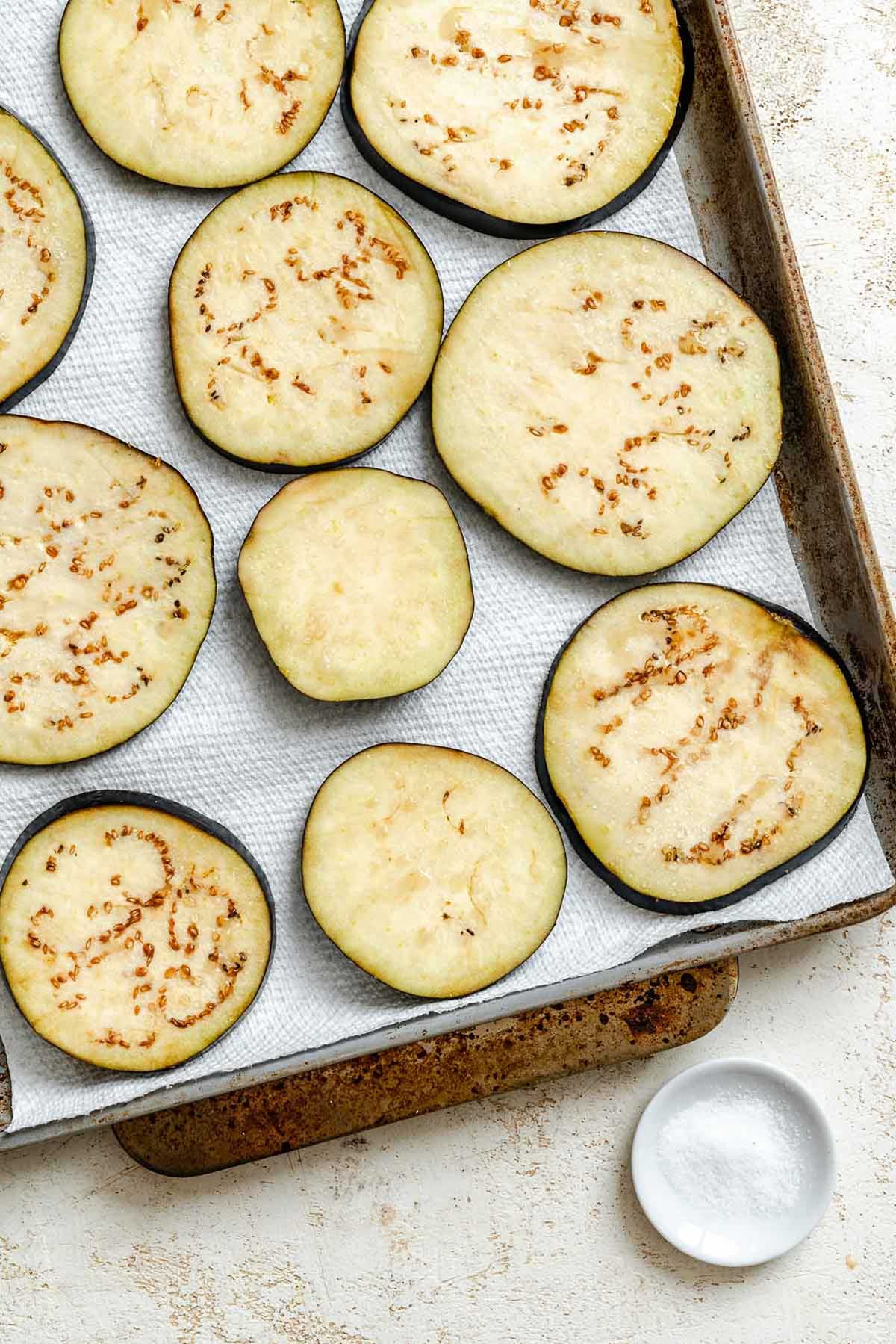 Thick slices of raw eggplant arranged on a paper towel-lined baking sheet, showing beads of moisture being drawn out by salt.