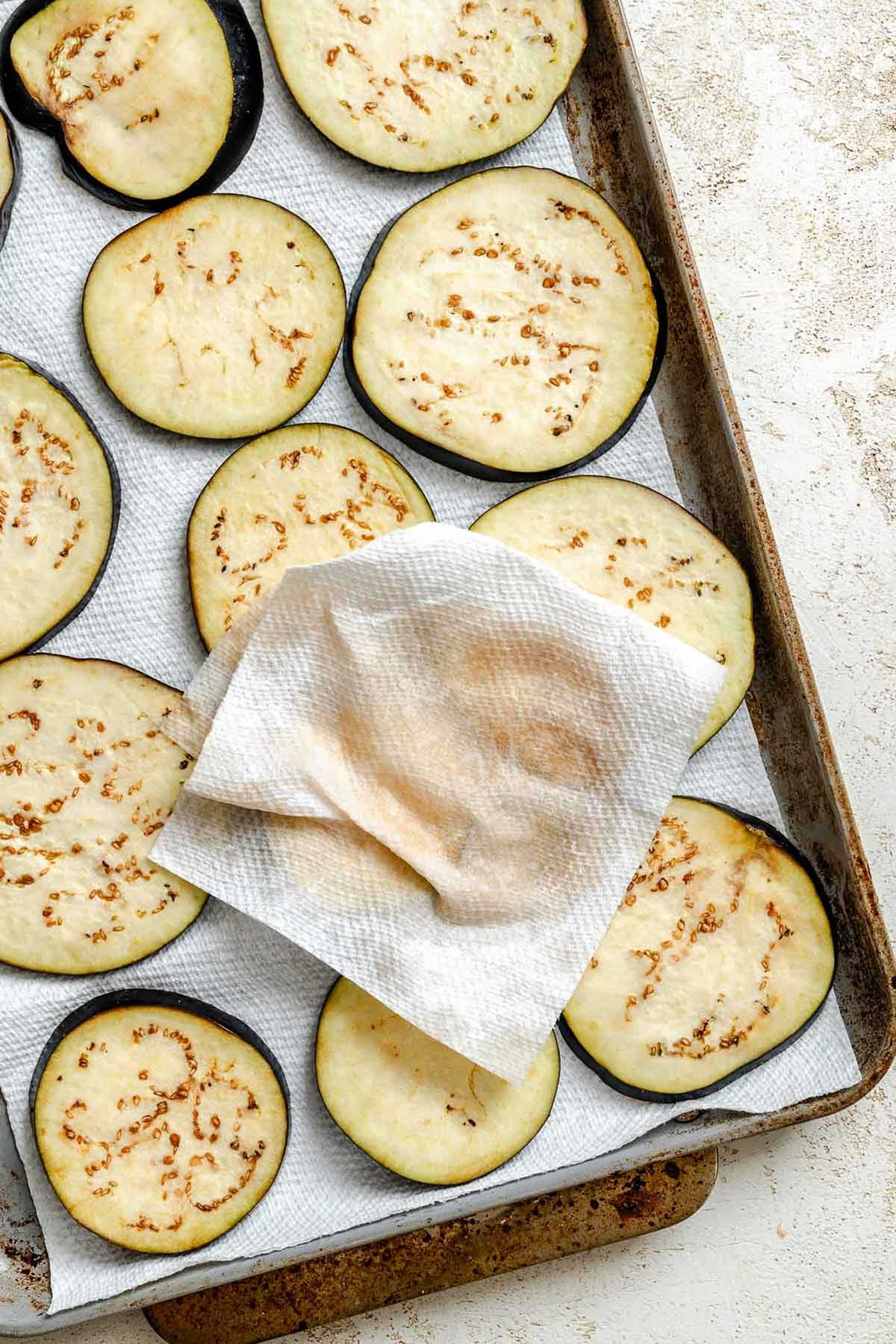 Eggplant slices on a baking sheet with a paper towel pressed on top to blot out excess moisture before cooking.