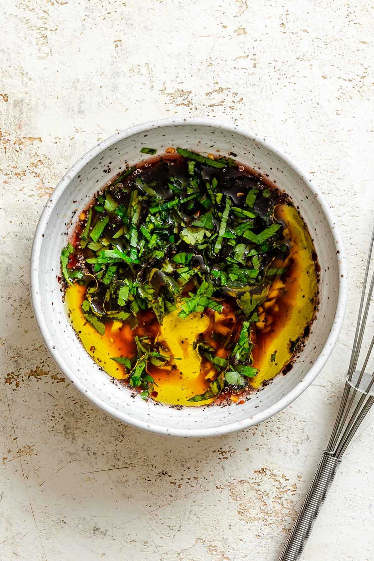 A white speckled bowl filled with a marinade of red wine vinegar, olive oil, chopped garlic, and fresh herbs, ready to be whisked.