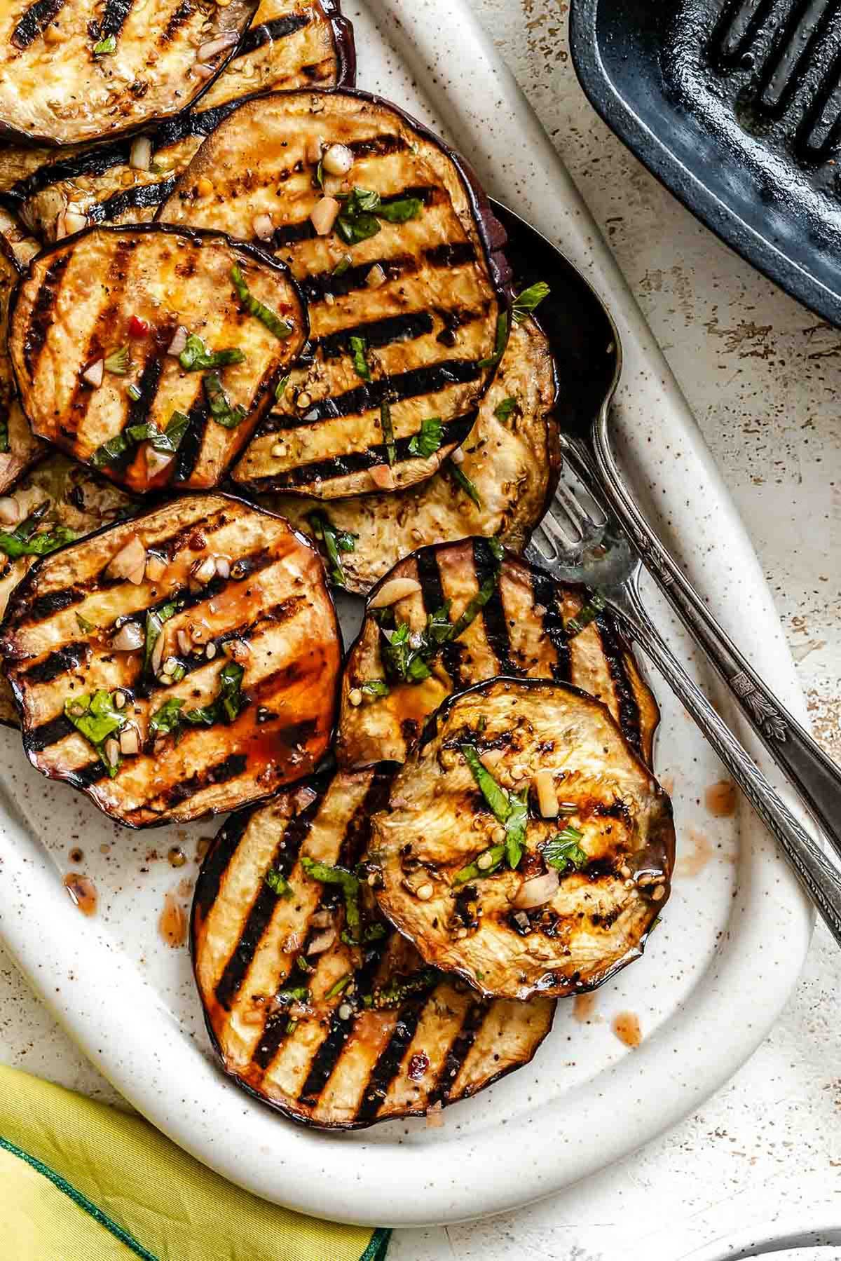A close-up of grilled eggplant slices on a white oval platter, garnished with chopped herbs and garlic from the marinade.