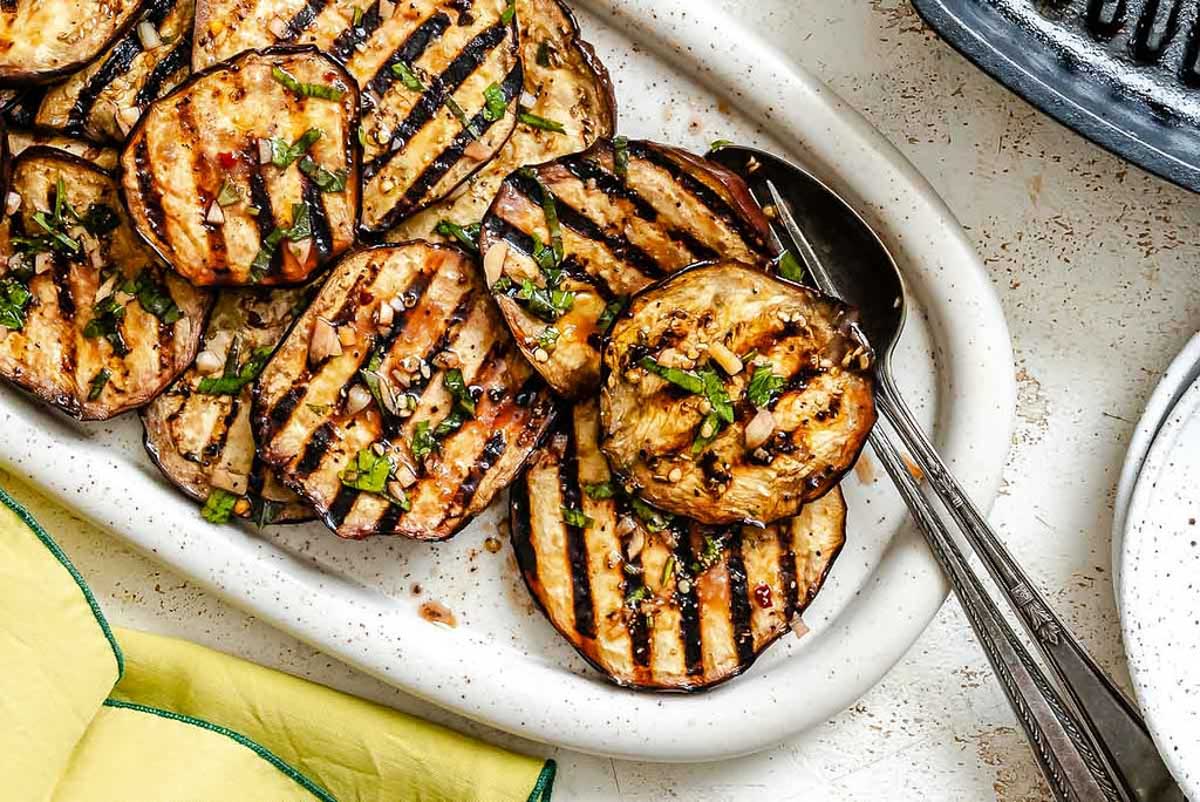 Overhead view of grilled and marinated eggplant slices served on a white platter with serving utensils.