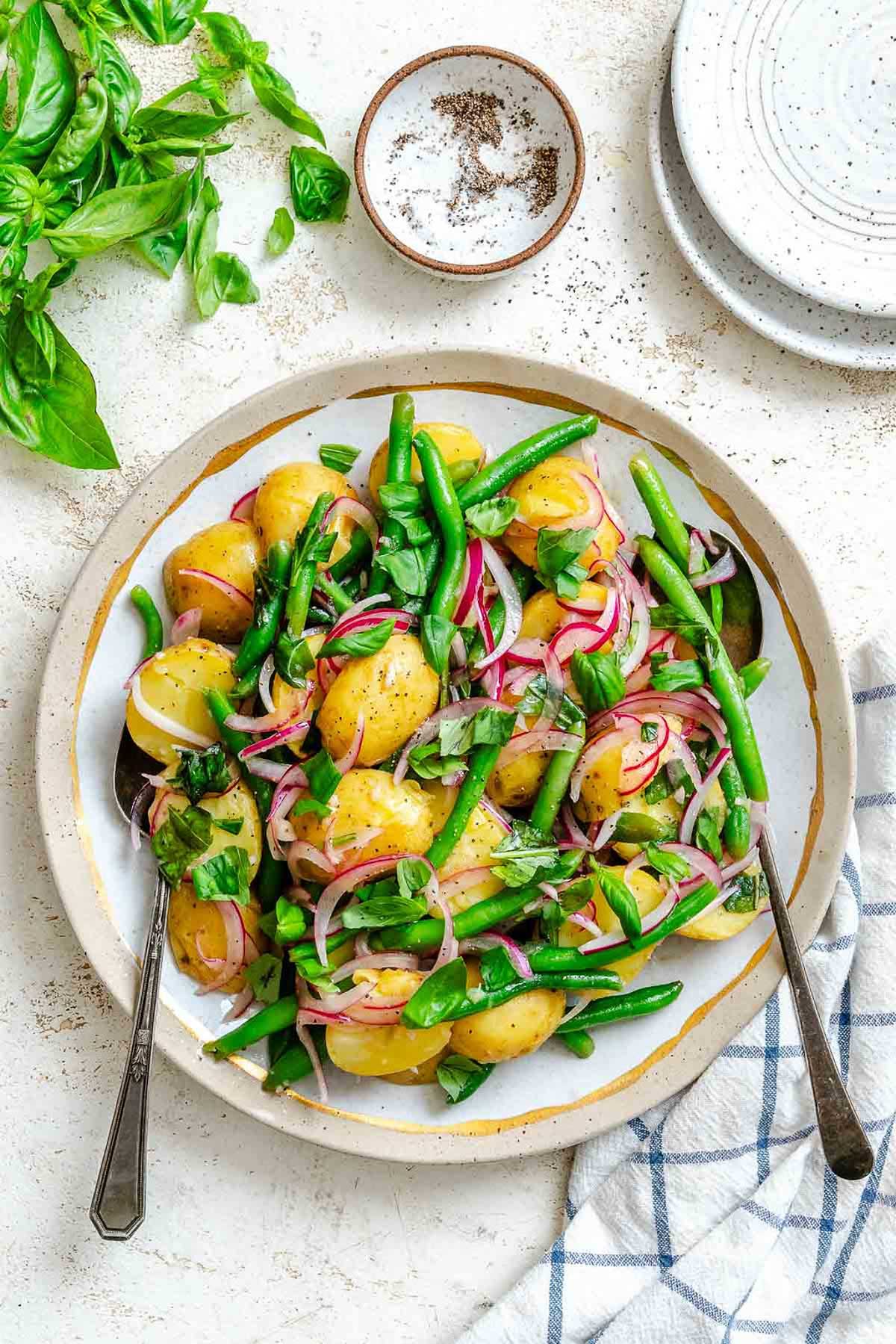 A bowl of Italian potato salad with halved yellow potatoes, green beans, red onions, and fresh basil leaves, lightly dressed and served with two spoons.