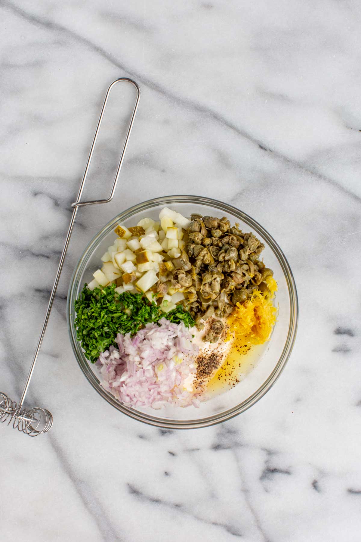 A glass mixing bowl on a marble surface filled with chopped ingredients for tartar sauce, including dill pickles, shallots, capers, parsley, lemon zest, and mayonnaise, arranged in neat sections before mixing. A metal whisk rests beside the bowl.