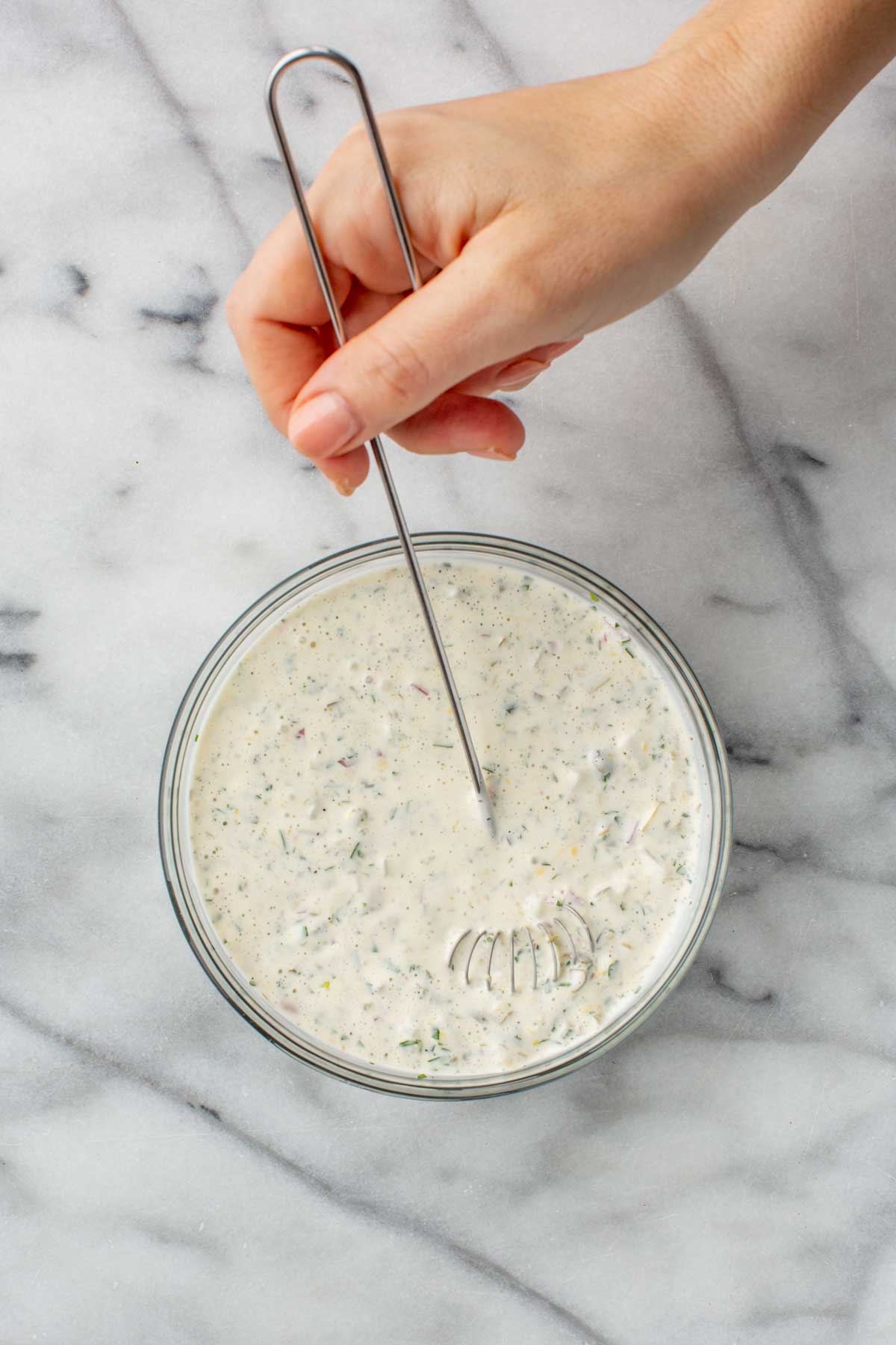 A hand holding a whisk stirs tartar sauce in a glass bowl, blending creamy mayonnaise with visible bits of parsley, shallots, and pickles. The bowl rests on a marble countertop.