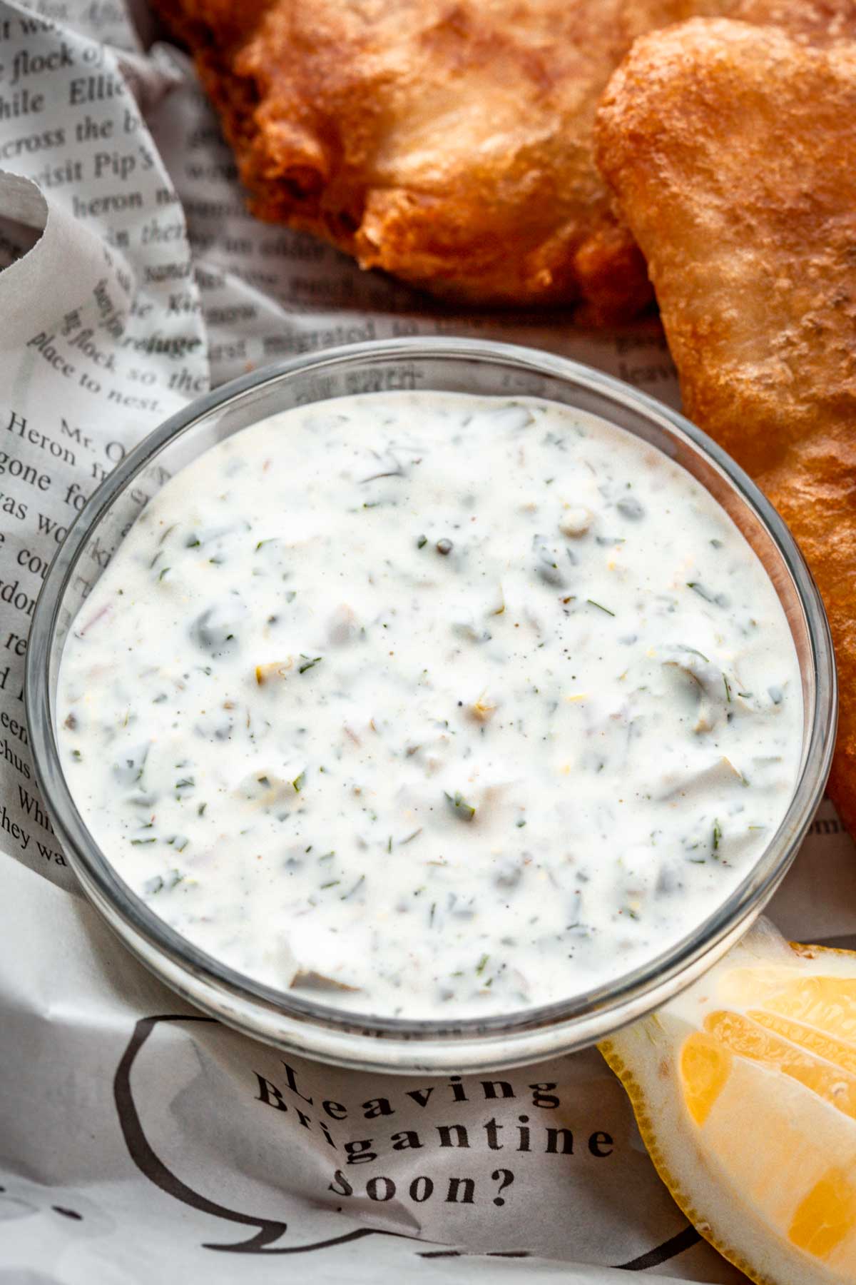 A small glass bowl filled with creamy tartar sauce, speckled with chopped herbs and pickles, placed next to crispy golden fried fish on newspaper-lined parchment.