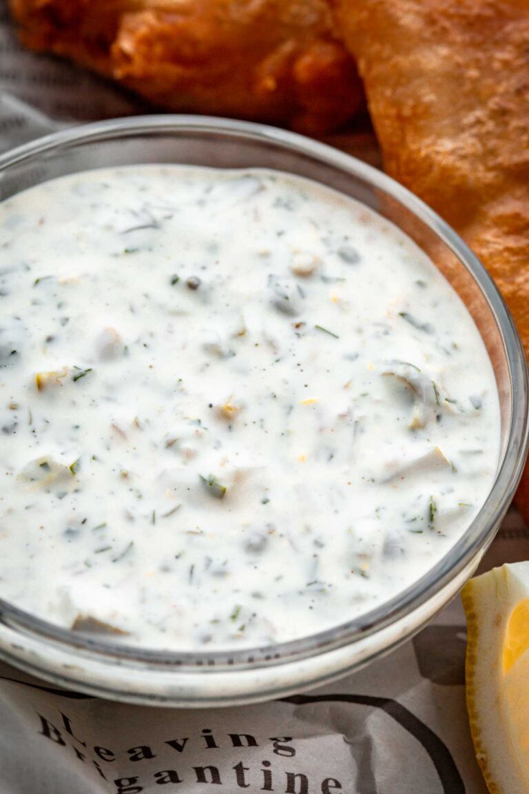 A close-up of thick tartar sauce in a bowl, showing finely chopped herbs, capers, and pickles suspended in a smooth, creamy base. Golden fried fish is partially visible in the background.