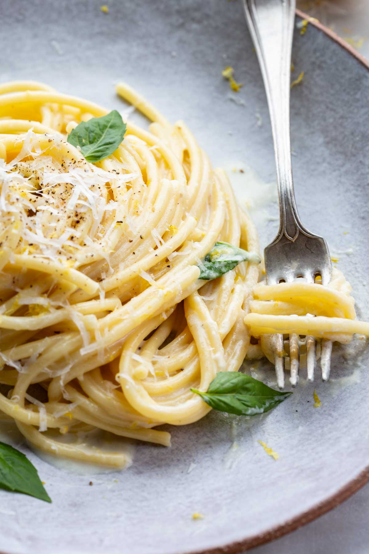 Close-up of pasta al limone with a fork twirling strands of spaghetti, garnished with basil and lemon zest on a stoneware plate.