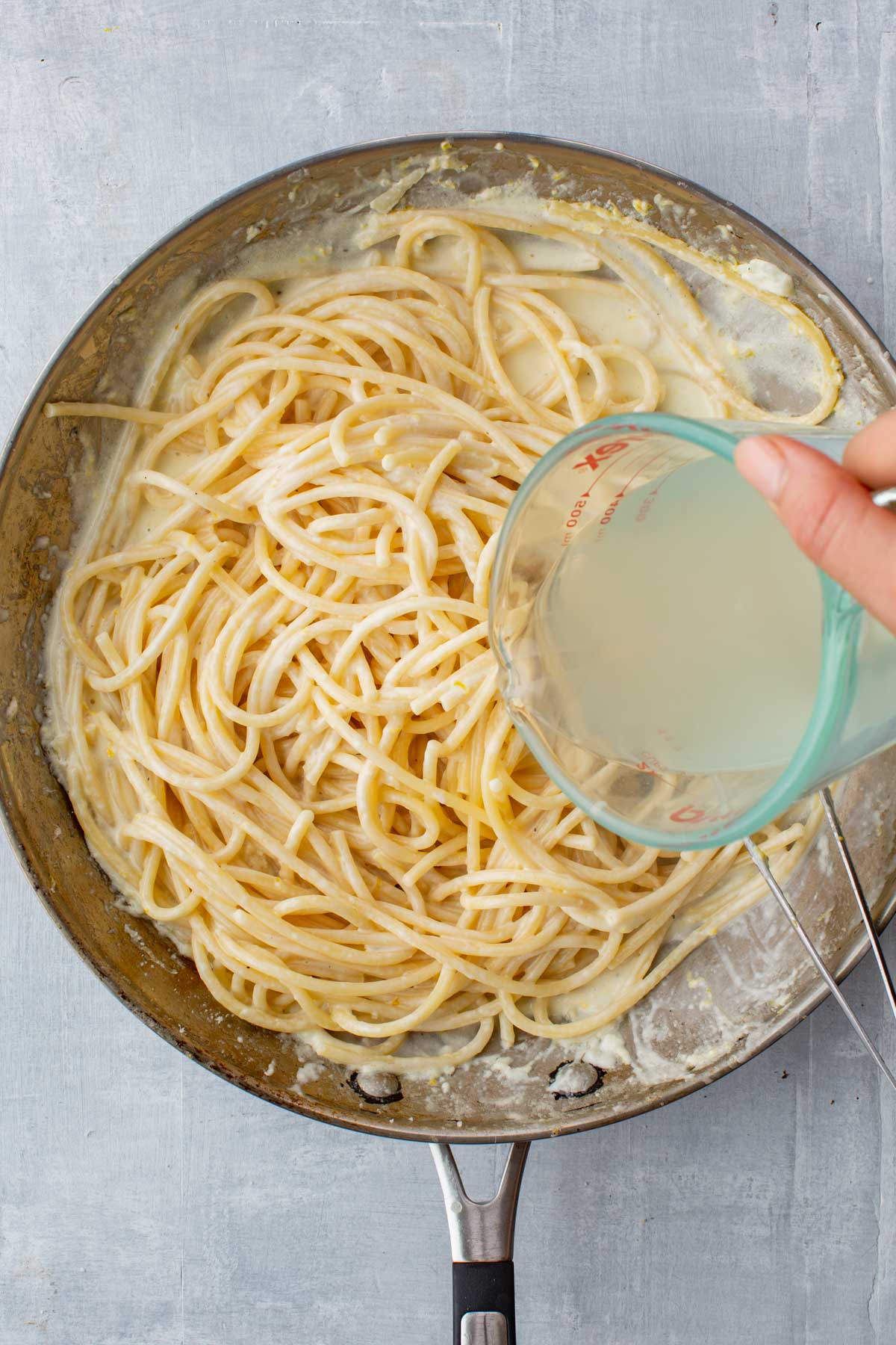 A hand pouring starchy pasta water from a glass measuring cup into a pan of spaghetti in lemon cream sauce to emulsify and thicken.