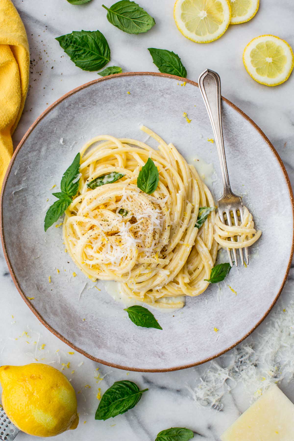 Top-down view of creamy pasta al limone on a ceramic plate with a fork and scattered basil leaves, Parmesan, and lemon rounds.