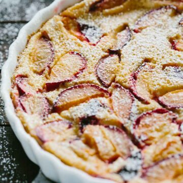 Close-up of a freshly baked plum clafoutis in a white tart dish, dusted with powdered sugar and filled with slices of ripe red plums.