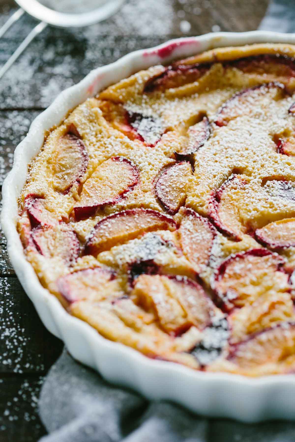 Close-up of a freshly baked plum clafoutis in a white tart dish, dusted with powdered sugar and filled with slices of ripe red plums.