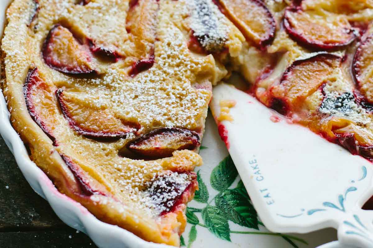 Side view of the plum clafoutis with one slice removed, revealing the custardy interior; a ceramic pie server stamped with “Eat Cake” is partially tucked underneath.