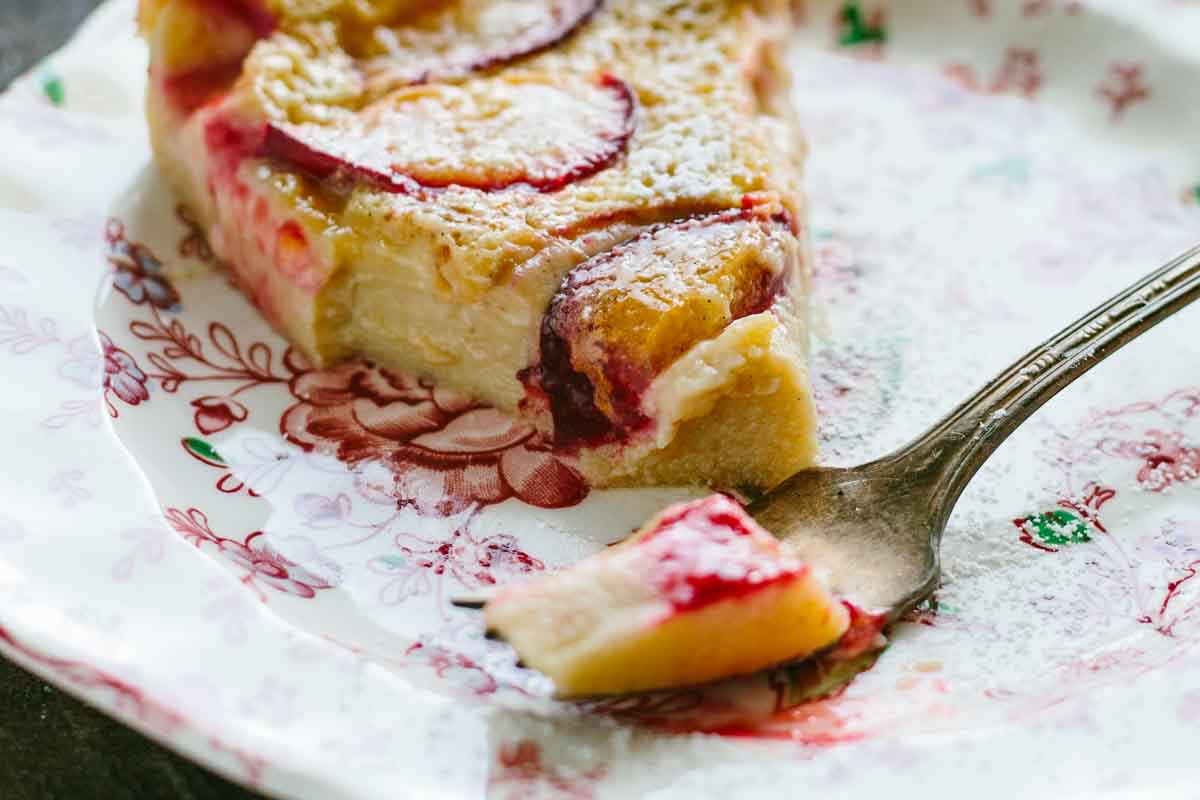 Individual slice of plum clafoutis on a vintage floral dessert plate, with a bite taken on a fork to show the soft texture and juicy plum pieces.