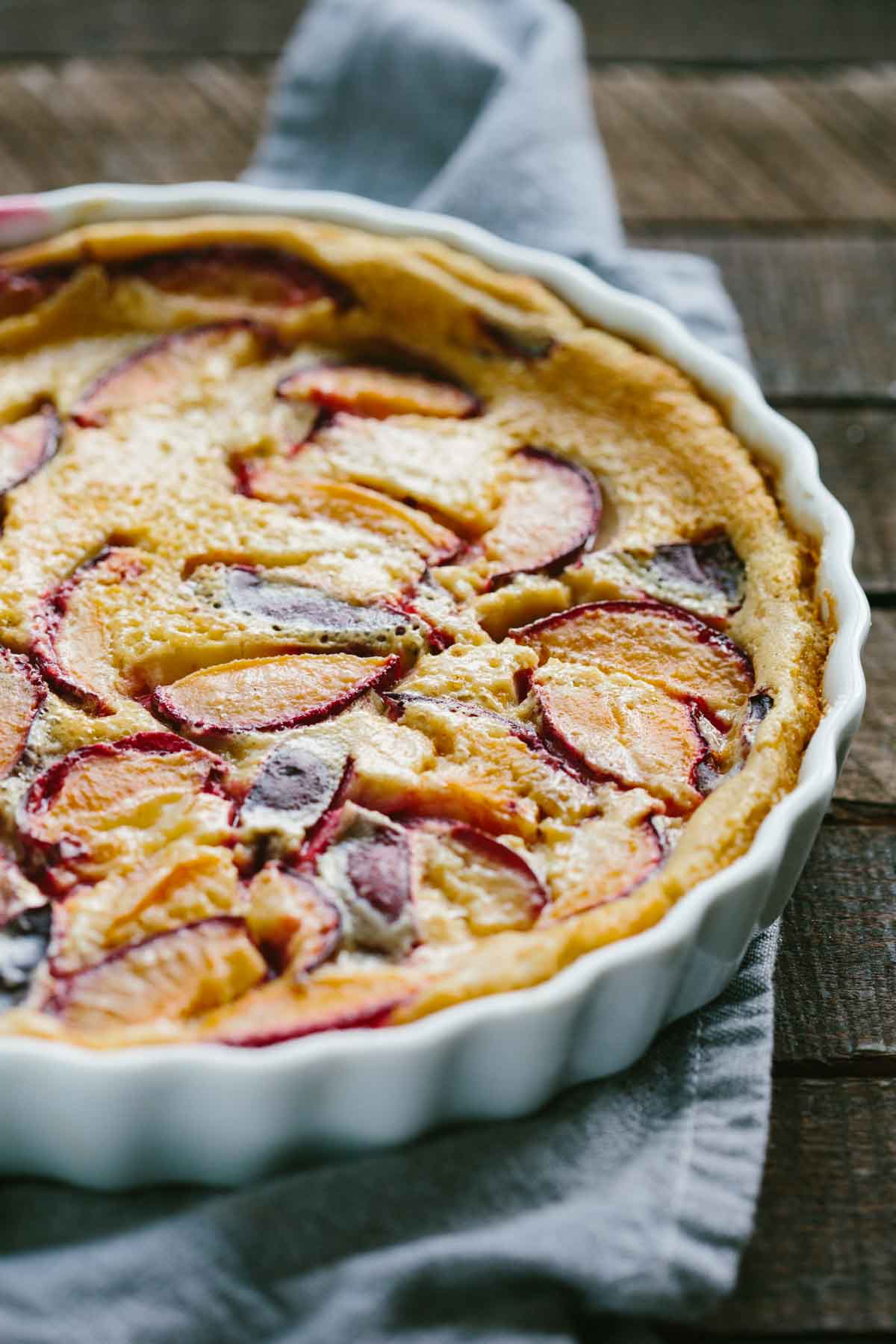 Side angle of the whole plum clafoutis in a fluted white dish set on a rustic wooden surface, showing golden baked edges and plum slices nestled in the creamy filling.