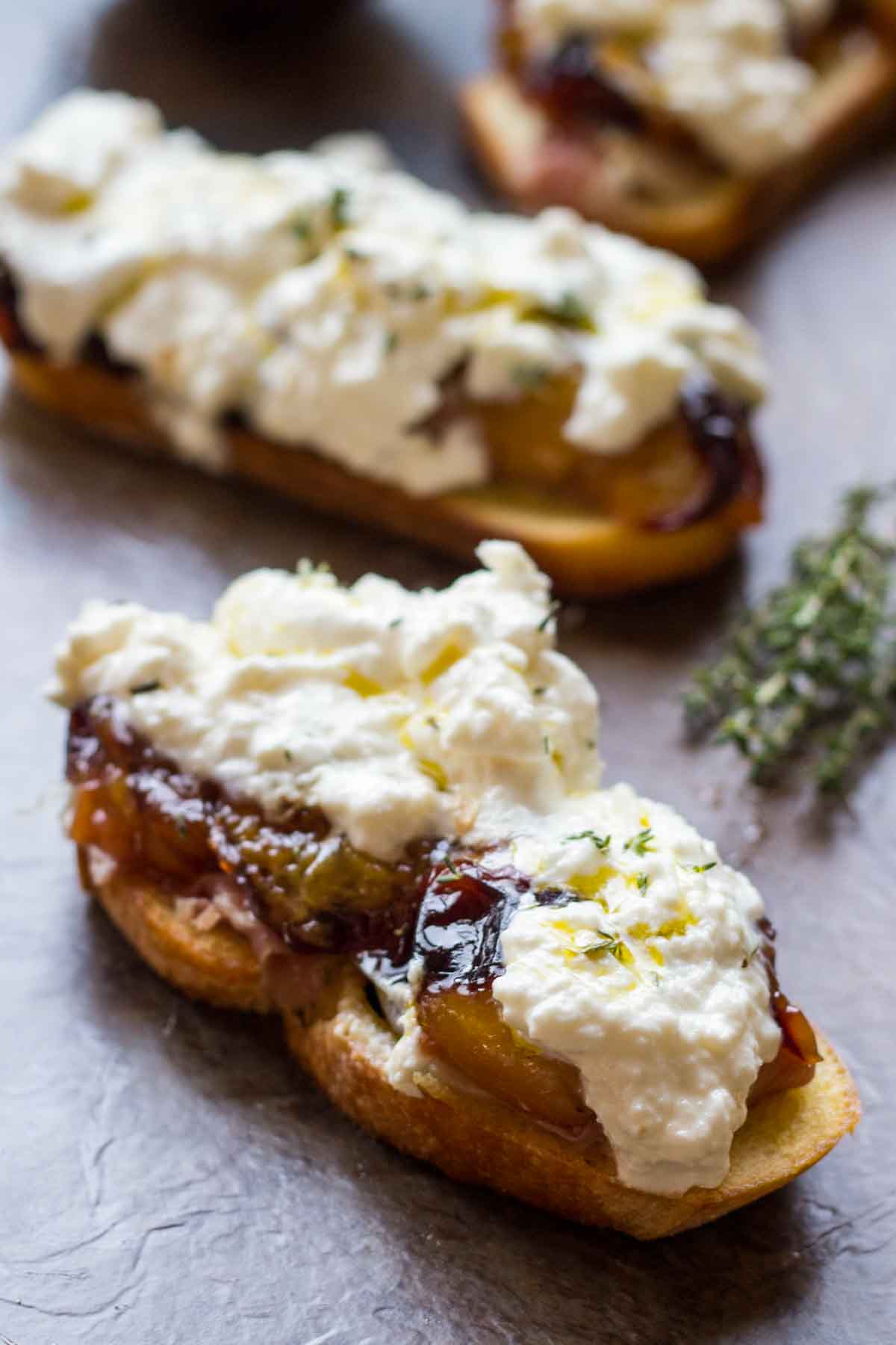 Close-up of crostini topped with roasted plums, ribbons of prosciutto, and a generous scoop of creamy burrata, garnished with thyme and olive oil