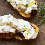 Top-down view of toasted crostini layered with jammy roasted plums, prosciutto, and fluffy burrata, sprinkled with fresh thyme.