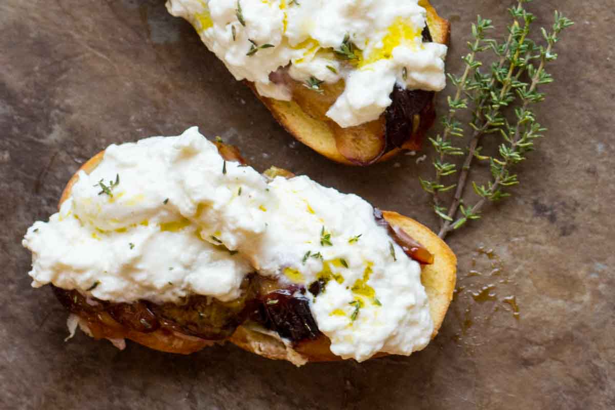 Top-down view of toasted crostini layered with jammy roasted plums, prosciutto, and fluffy burrata, sprinkled with fresh thyme.