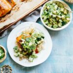 Colorful table scene with a serving of glazed salmon over rice and cucumber avocado salad surrounded by a baking tray of salmon, bowls of rice and salad, and scattered sesame seeds.