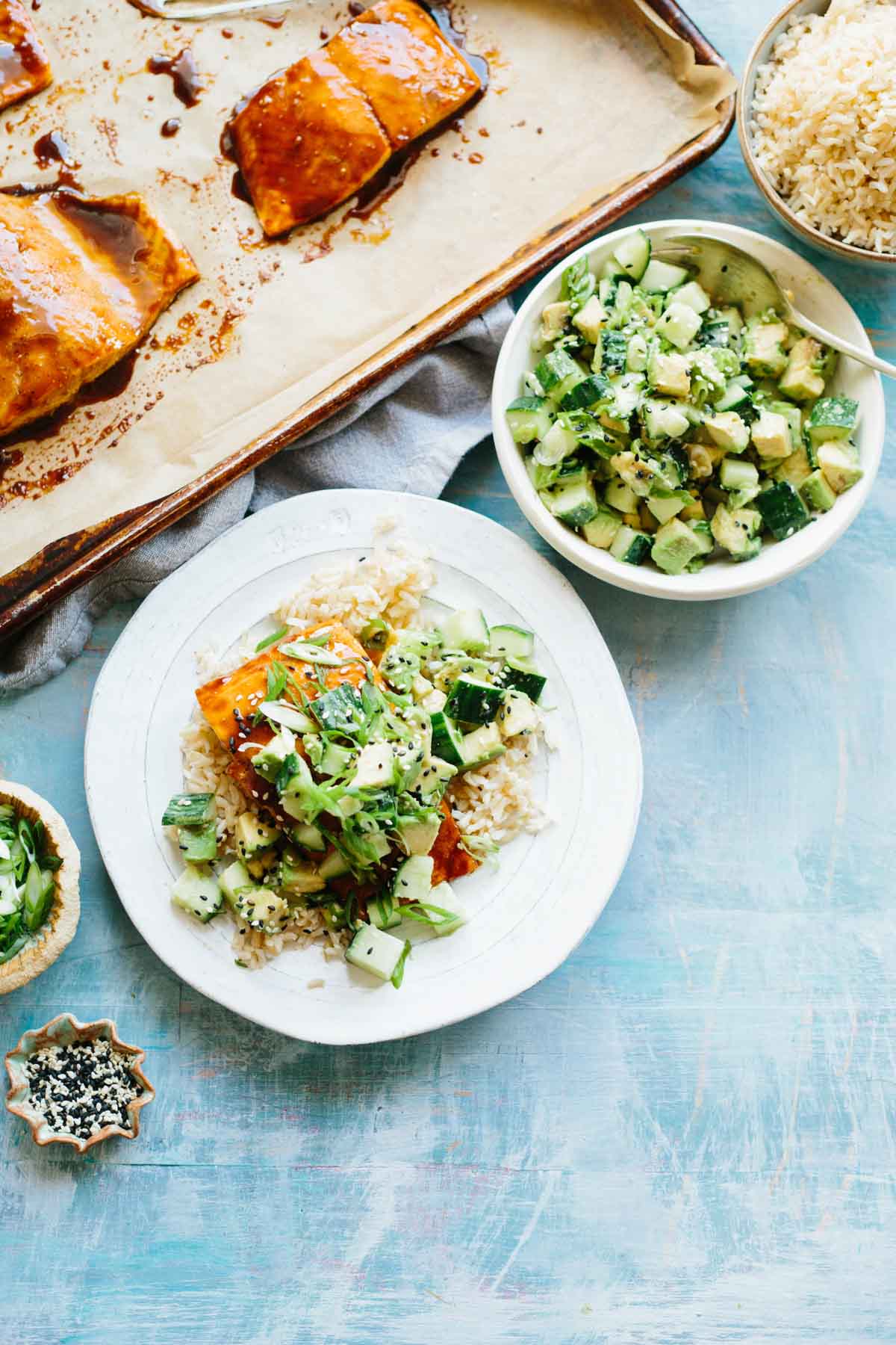Colorful table scene with a serving of glazed salmon over rice and cucumber avocado salad surrounded by a baking tray of salmon, bowls of rice and salad, and scattered sesame seeds.