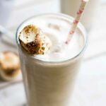 Side view of a toasted marshmallow milkshake in a tall clear glass with a pink-striped straw and a perfectly browned marshmallow garnish.
