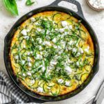 Overhead view of a golden-brown zucchini frittata in a cast iron skillet, topped with crumbled feta cheese and fresh basil ribbons.