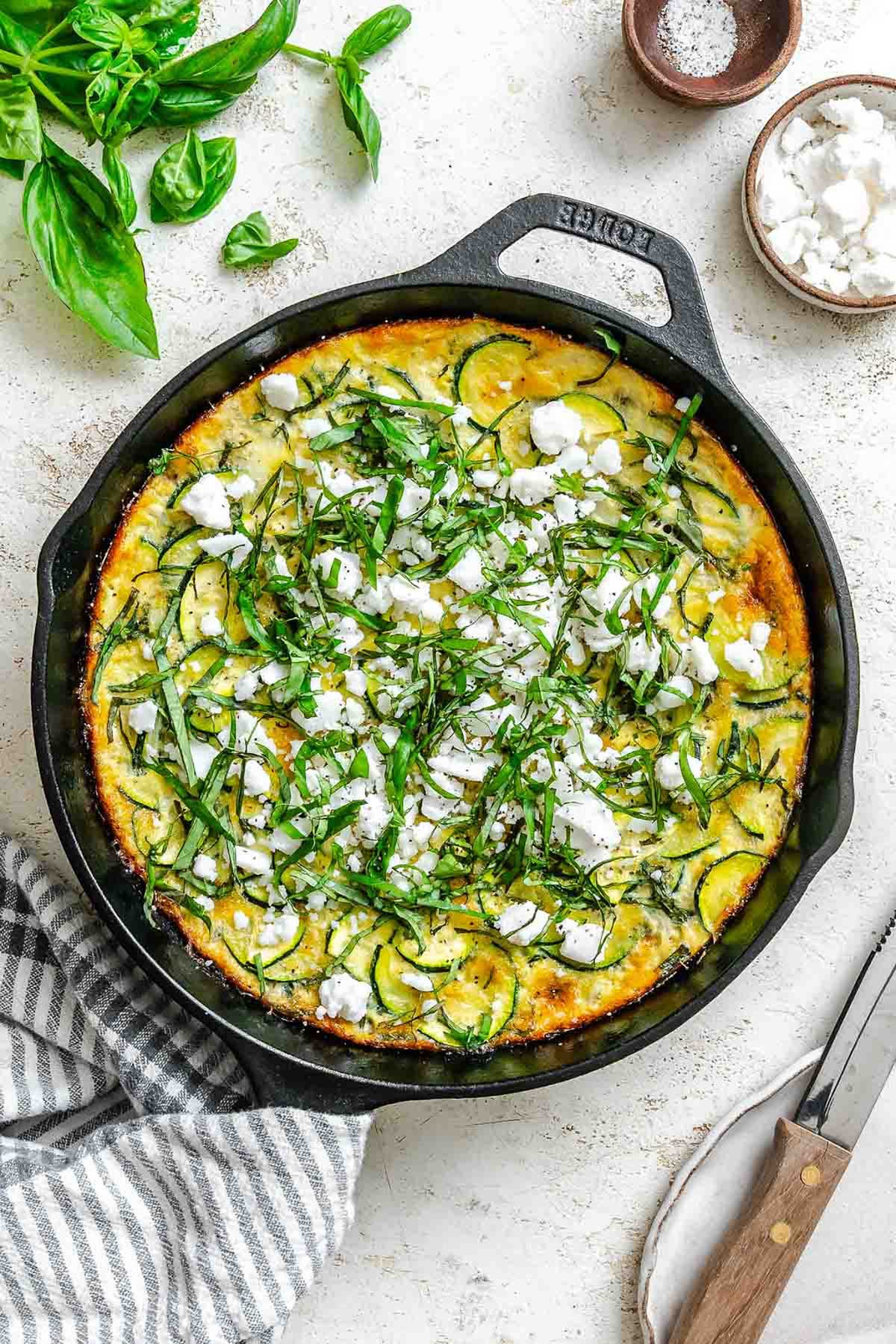 Overhead view of a golden-brown zucchini frittata in a cast iron skillet, topped with crumbled feta cheese and fresh basil ribbons.