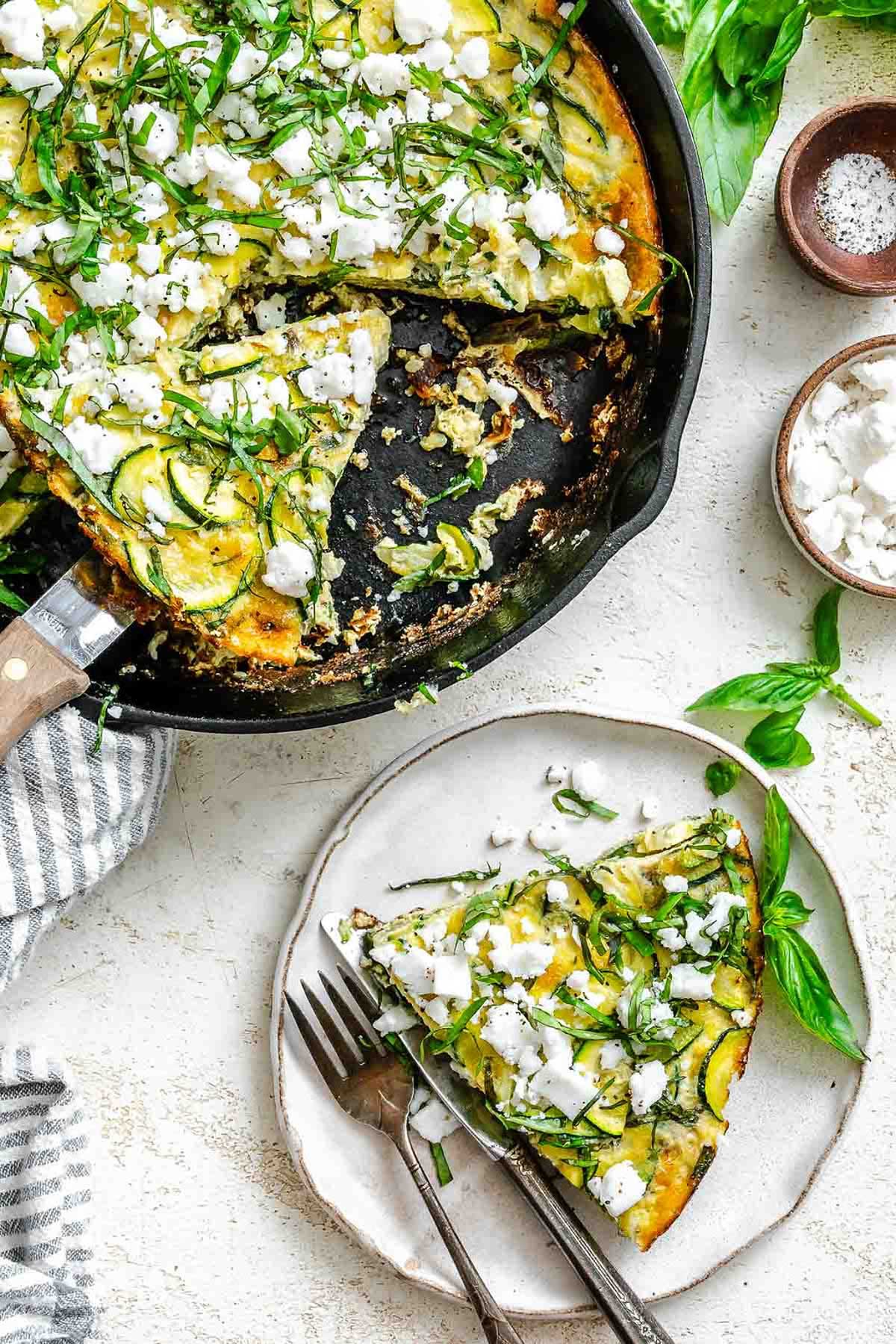 A slice of zucchini frittata on a plate beside the skillet, garnished with fresh basil and feta, ready to be served with forks.