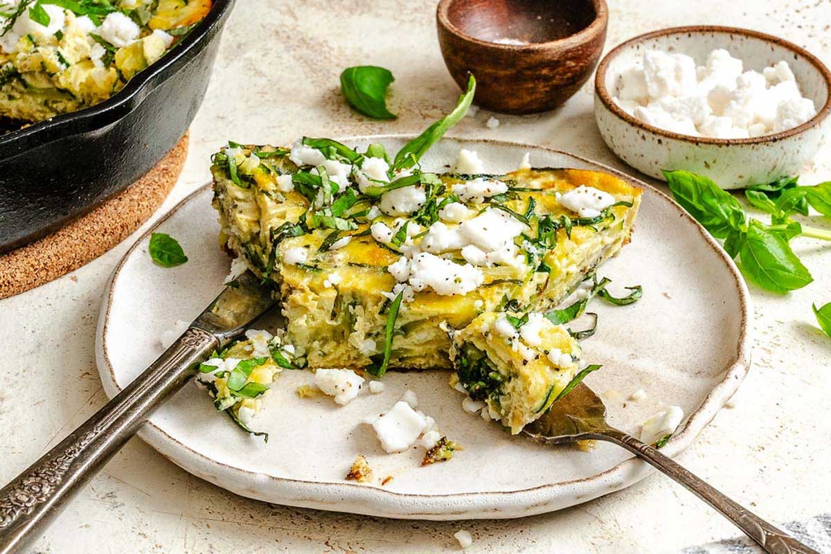 Side view of a thick slice of zucchini frittata on a ceramic plate, showing its layered interior and crumbled feta on top.