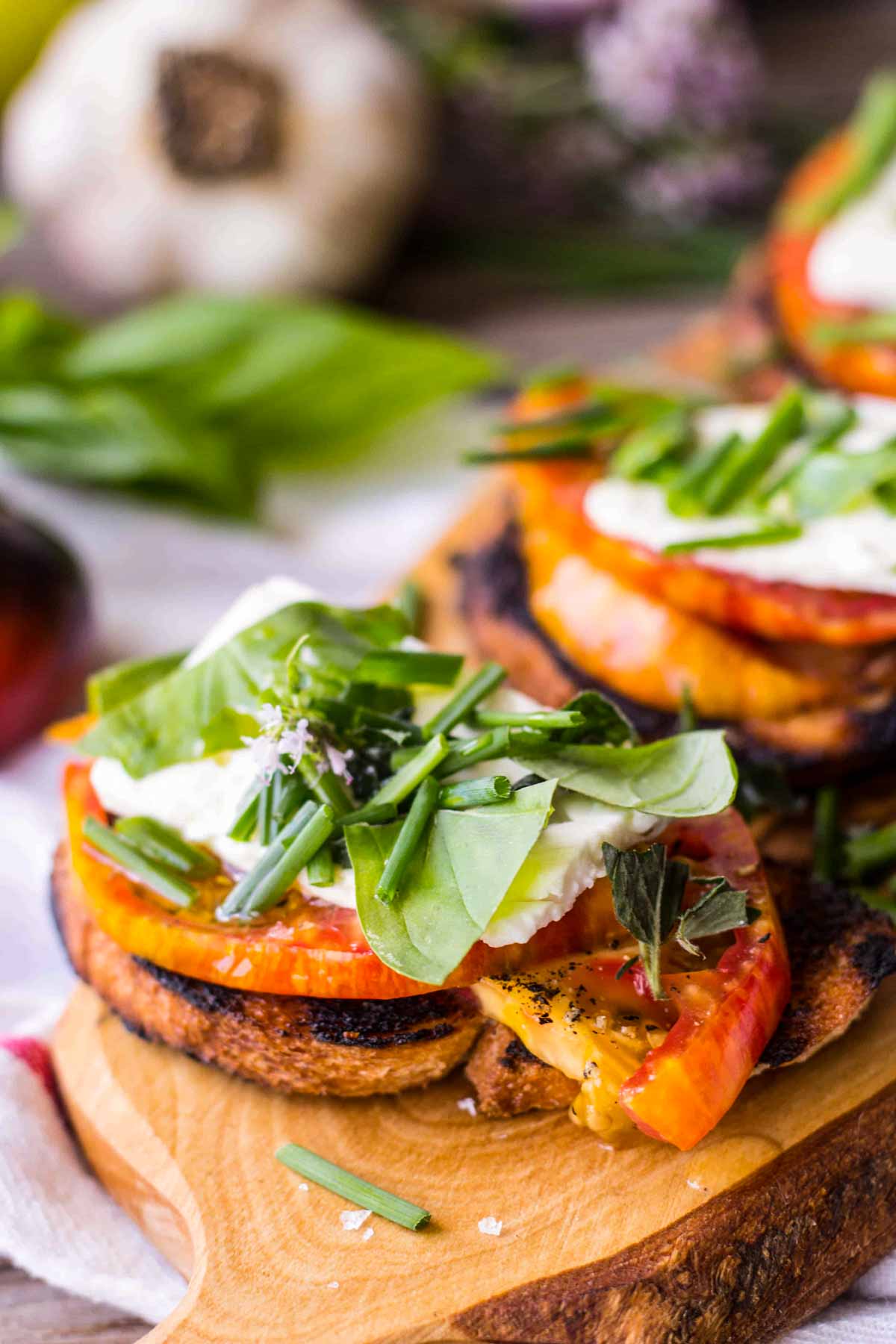 Close-up of a grilled toast layered with thick heirloom tomato slices, a dollop of creamy cheese, and a generous scatter of fresh basil, chives, and oregano on a wooden board.