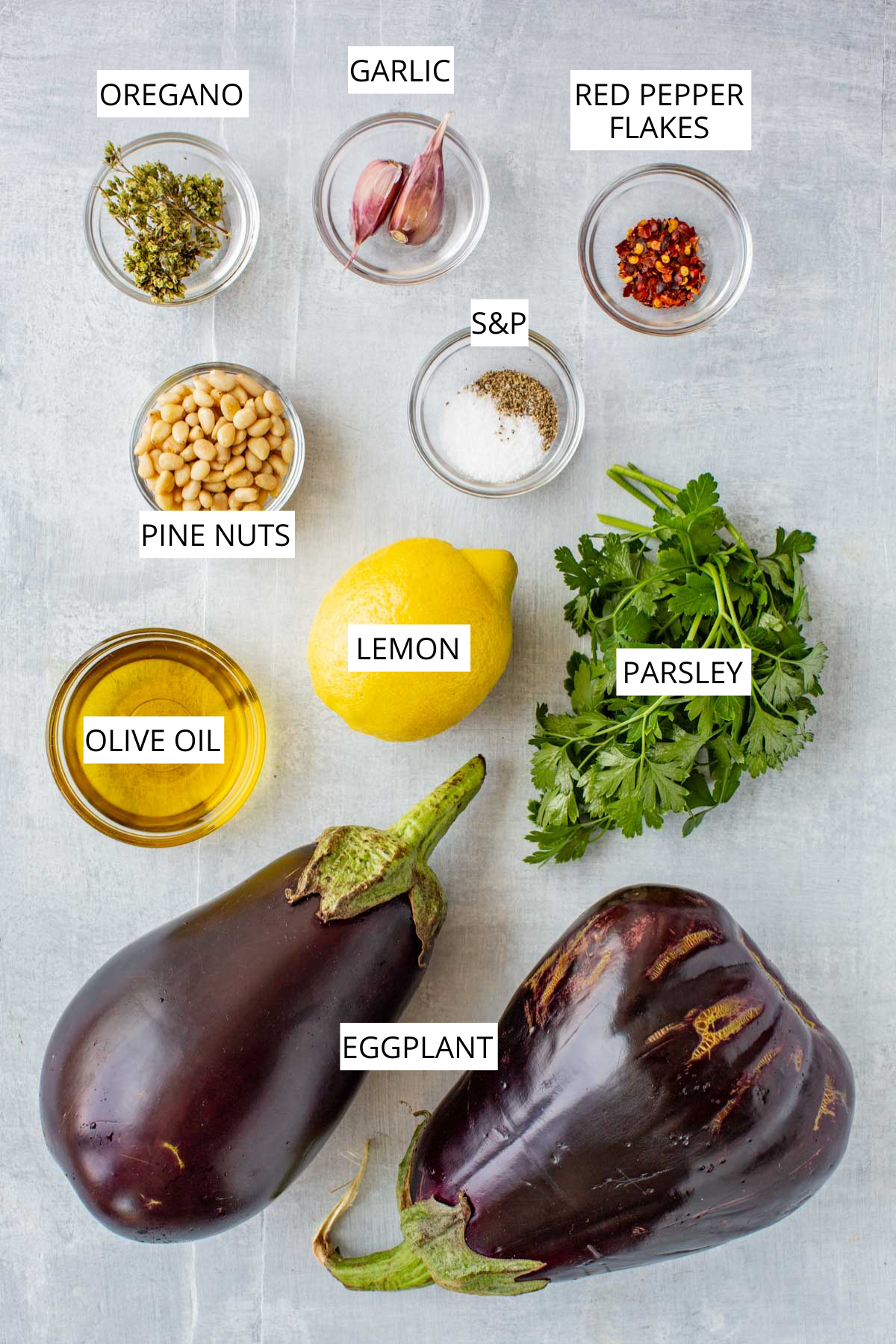 Charred eggplant dip ingredients - Overhead view of labeled ingredients including whole eggplants, garlic cloves, fresh parsley, oregano, red pepper flakes, pine nuts, lemon, salt and pepper, and olive oil.