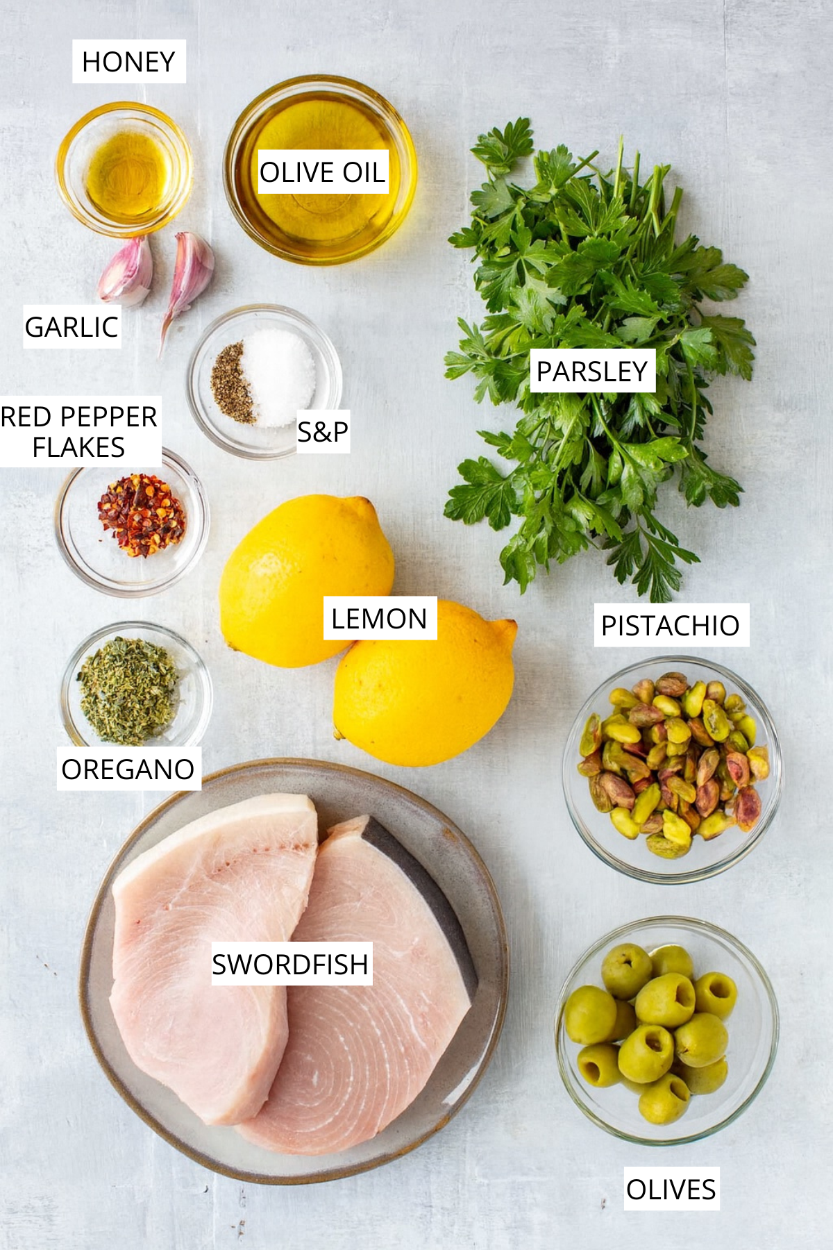 Overhead photo of labeled ingredients including swordfish steaks, olive oil, lemon, garlic, parsley, oregano, red pepper flakes, pistachios, and Castelvetrano olives on a gray surface.