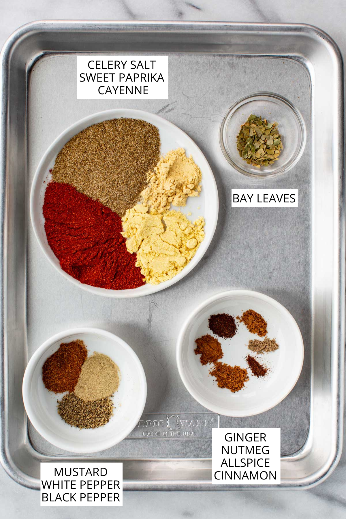 Overhead view of individual spices labeled for homemade Old Bay seasoning, including celery salt, paprika, cayenne, mustard, peppers, bay leaves, and warm spices arranged neatly on a silver baking sheet.