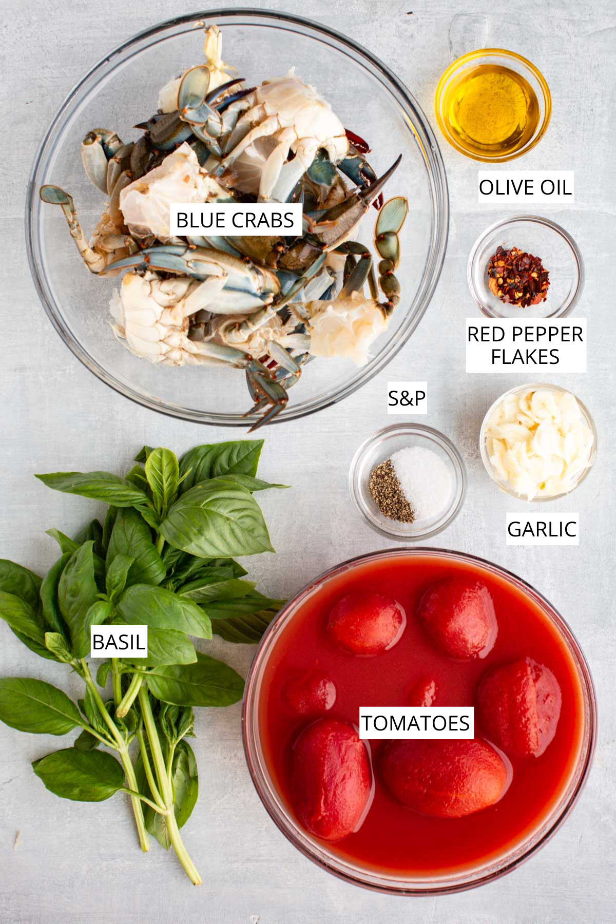 Overhead shot of raw recipe ingredients labeled: fresh blue crabs, olive oil, red pepper flakes, salt and pepper, garlic, fresh basil, and whole canned tomatoes in juice.