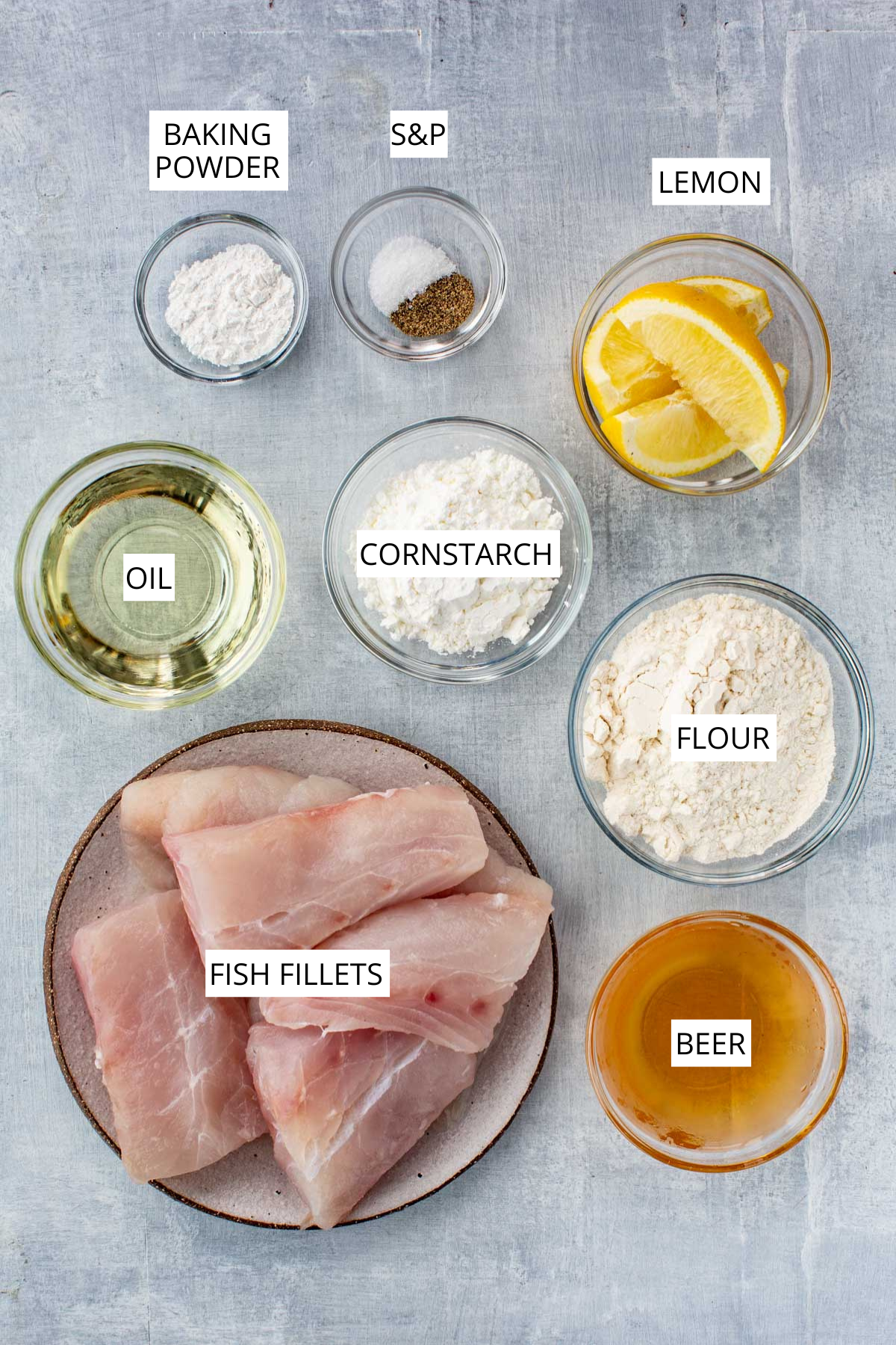 Overhead view of labeled beer-battered fish ingredients, including fish fillets, flour, cornstarch, beer, oil, lemon wedges, baking powder, salt, and pepper.