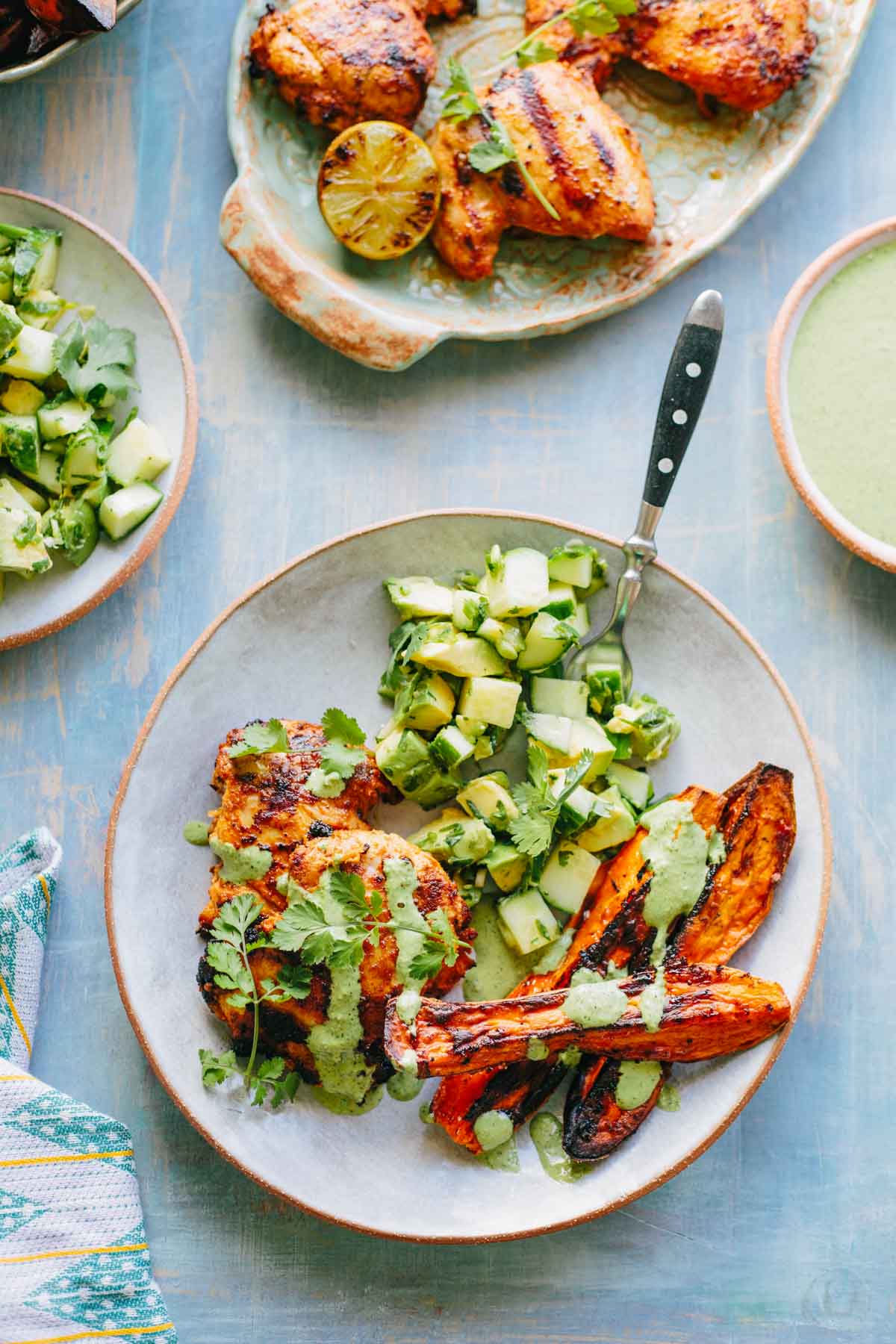 Peruvian grilled chicken with avocado salad and roasted sweet potatoes - A plate with charred chicken thighs drizzled in creamy green sauce, served alongside roasted sweet potato halves and a fresh cucumber avocado salad, garnished with cilantro.