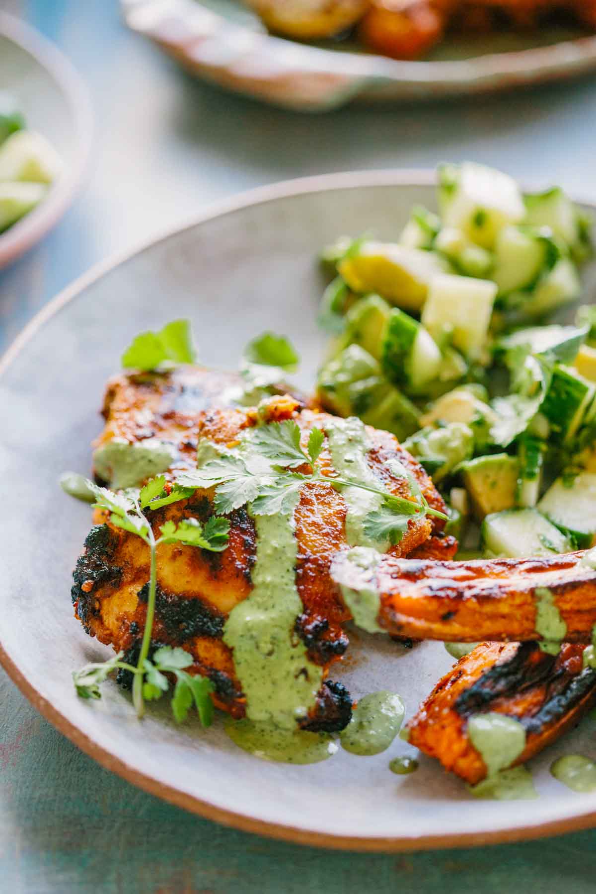 Close-up of grilled chicken with green sauce - Juicy chicken thighs topped with herbaceous Peruvian green sauce, fresh cilantro leaves, and smoky roasted sweet potatoes on the side.