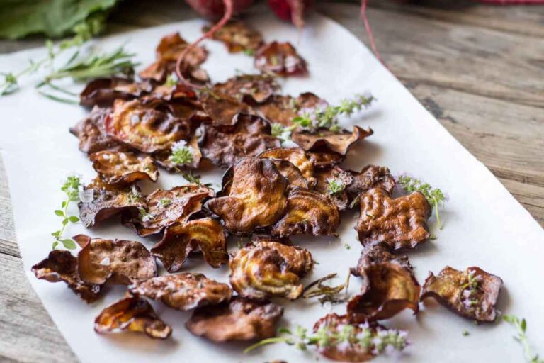 Rustic beet chips on parchment – Crispy oven-roasted beet chips scattered over parchment paper and garnished with fresh herbs on a wooden surface.