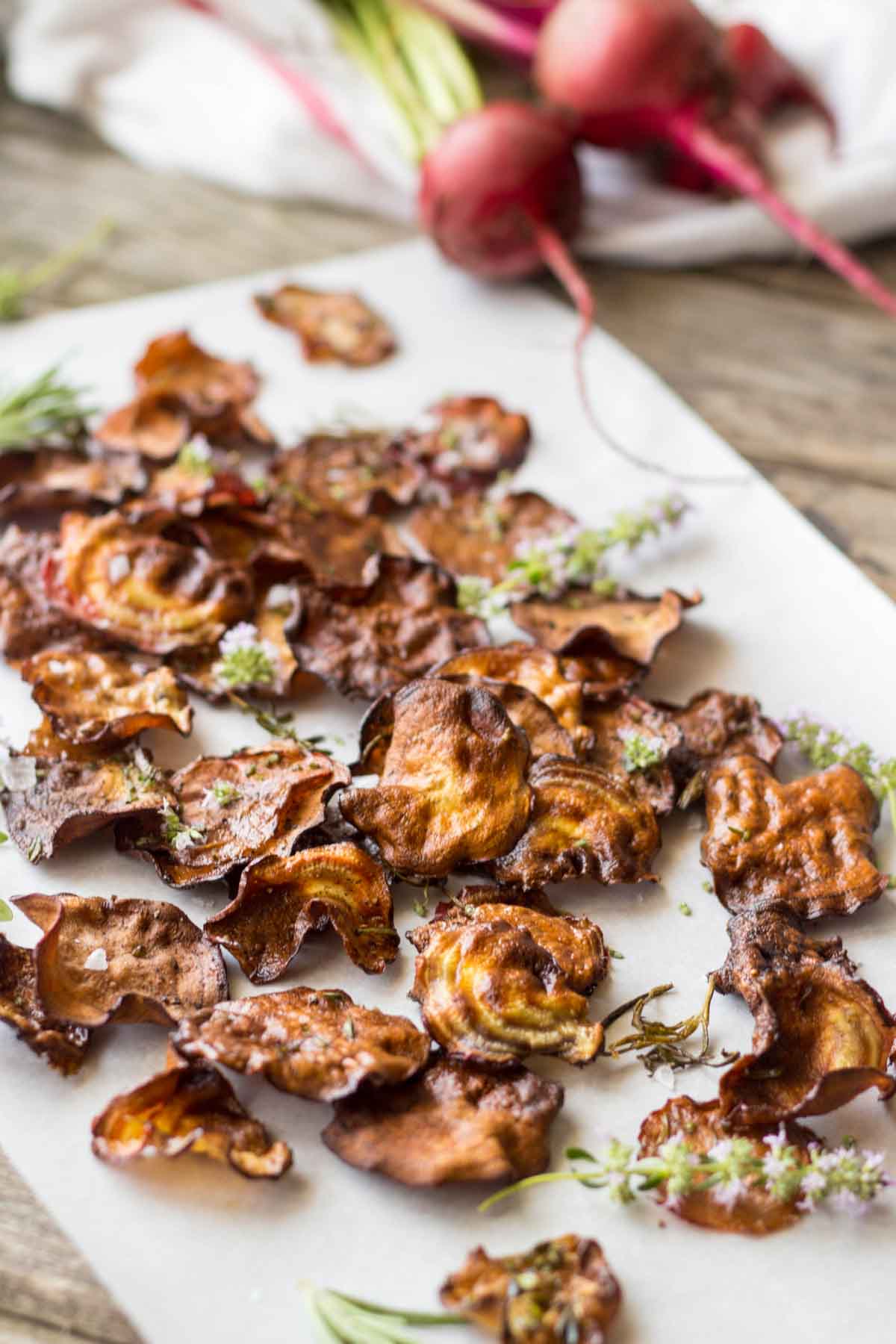 Close-up of beet chips with fresh beets - Golden and crimson beet chips on parchment paper with raw beets and leafy tops blurred in the background, sprinkled with herbs and flaky salt.