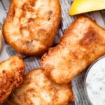 Crispy beer-battered fish on newspaper with lemon wedges and a small bowl of tartar sauce.