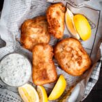 Overhead view of beer-battered fish served on newspaper with lemon wedges and tartar sauce.