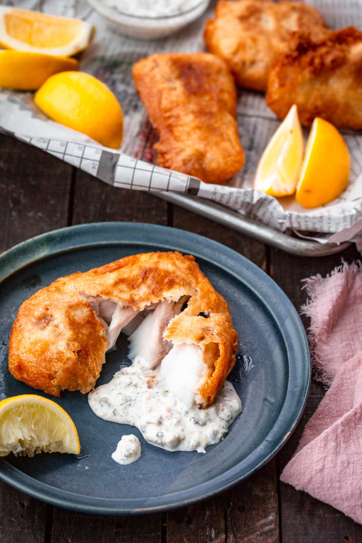 Plate with a piece of beer-battered fish broken open over tartar sauce, with lemon wedges on the side, and more fish in the background.