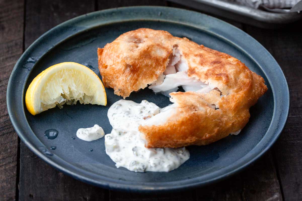 Close-up of beer-battered fish broken open to reveal flaky white flesh, served with tartar sauce and a lemon wedge on a dark plate.