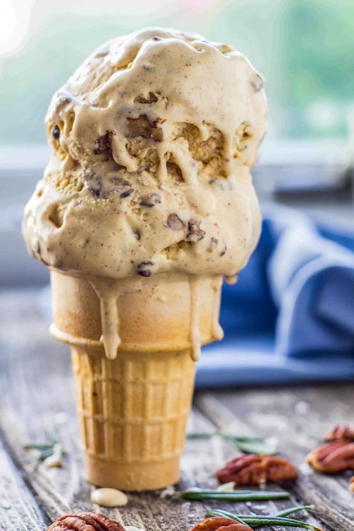 Two large scoops of rosemary brown butter pecan ice cream in a waffle cone, melting down the sides, with pecans and rosemary scattered on a rustic wooden table.