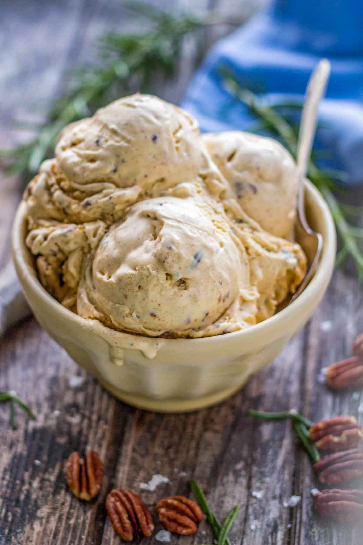 A cream-colored bowl filled with multiple scoops of rosemary brown butter pecan ice cream, surrounded by fresh rosemary sprigs and toasted pecans on a weathered wood surface.
