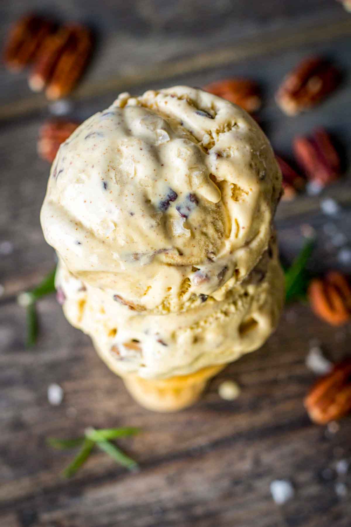 Overhead view of a triple-scoop rosemary brown butter pecan ice cream cone on a wooden surface, with coarse salt crystals, rosemary sprigs, and pecans scattered around.
