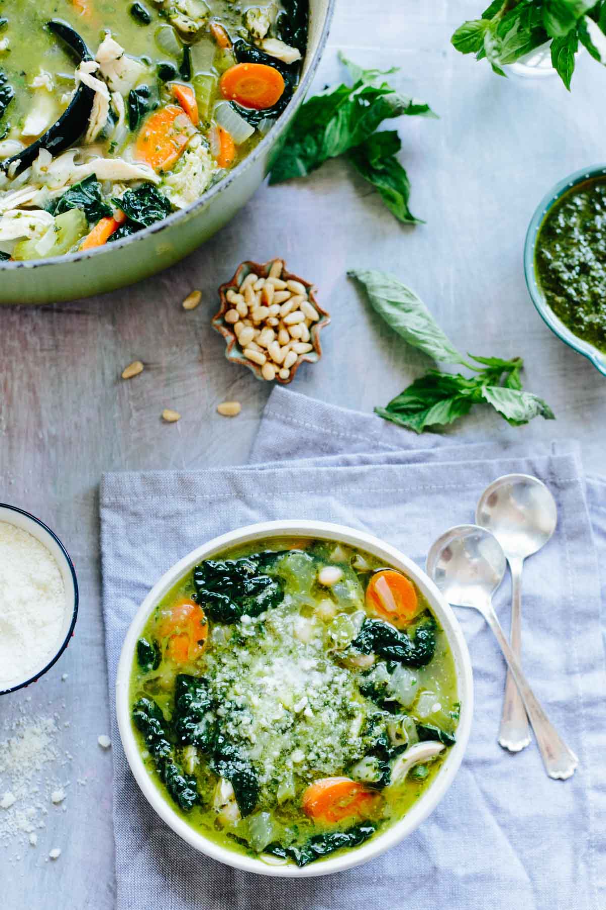 Overhead scene of a bowl of soup served next to the pot, surrounded by basil leaves, pine nuts, and parmesan cheese.