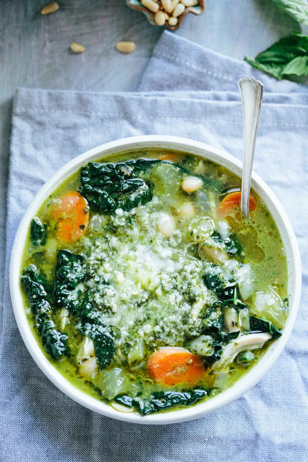 Overhead close-up of a white bowl filled with chicken, kale, and white bean soup topped with pesto and grated parmesan.