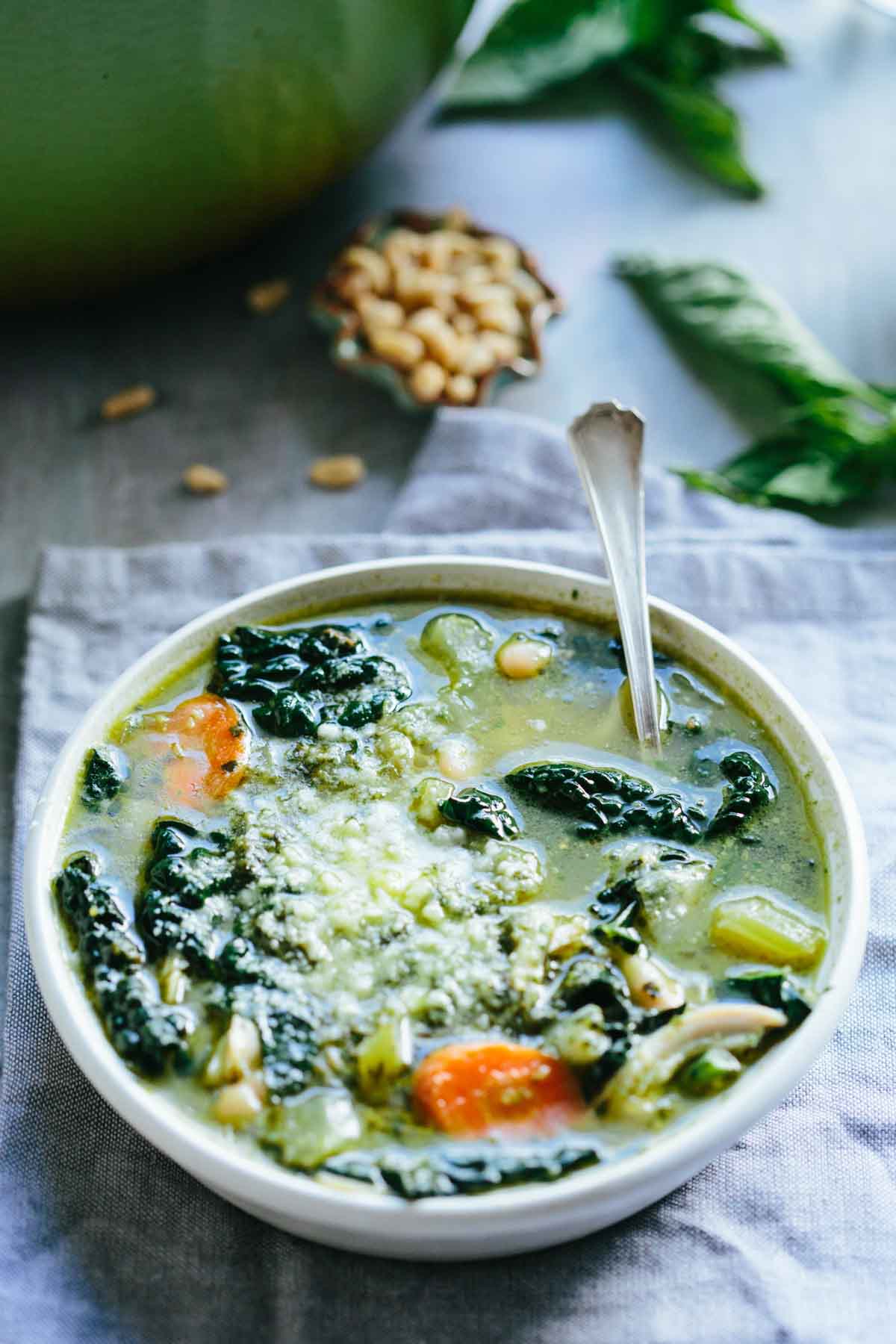 Side angle of a bowl of pesto chicken soup, showing carrots, kale, and beans under a layer of melted parmesan cheese.