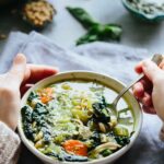 Hands holding a bowl of chicken pesto soup with a spoon dipping in, showing hearty vegetables and greens.