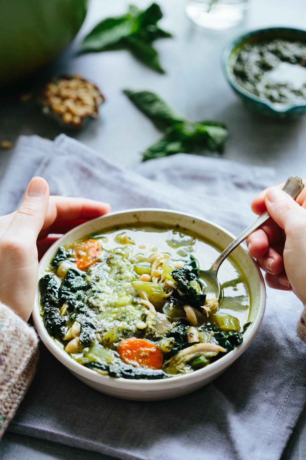 Hands holding a bowl of chicken pesto soup with a spoon dipping in, showing hearty vegetables and greens.